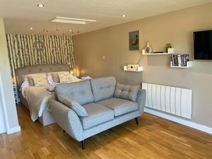 A modern bedroom with a bed, grey sofa, wooden flooring, a radiator, and a TV mounted on the wall. - Pheasant Lodge: Cozy Retreat (Wimborne)