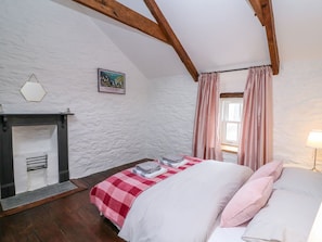 Ein Schlafzimmer mit Kamin, einem Bett mit rosa kariertem Überwurf, Holzbalken und einem Fenster mit Vorhängen.