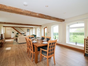 A spacious dining area with wooden flooring, a large window with a view of greenery, and a staircase leading to another room.