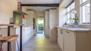 A narrow kitchen with white cabinets, a wooden table, and a window with a plant. - Field Head House, Hawkshead (Ulverston)