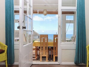 An open door leading to a balcony with a table and chairs, a view of the sea, and a hanging light fixture.