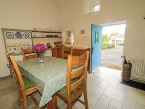 A dining area with a table, chairs, a vase with flowers, a kitchen in the background, and a blue door leading outside.