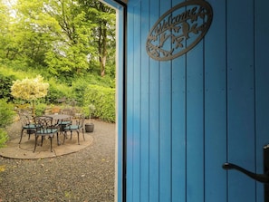 Puerta azul con un cartel que dice 'Welcome', área de estar exterior con sillas y una mesa, rodeada de follaje.