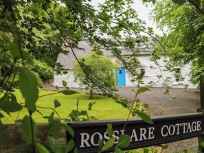 Una cabaña con puerta azul, rodeada de follaje, y un letrero que dice 'ROSS LARE COTTAGE'.