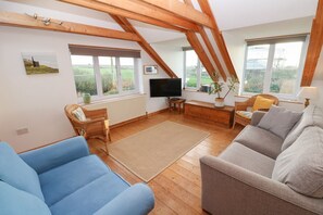 Ein Zimmer mit hoher Decke, Holzbalken, einem Fernseher, einer Couch, einem Sessel, einem kleinen Tisch und Blick ins Freie.