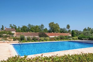 Ein rechteckiger Pool mit einer gefliesten Terrasse, umgeben von Grünflächen und Gebäuden mit roten Ziegeldächern.
