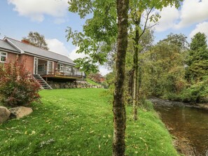 A house with a stone staircase leading to a deck, a grassy lawn, and a stream. - Ger Yr Afon Cottage, Rhayader (Rhayader)