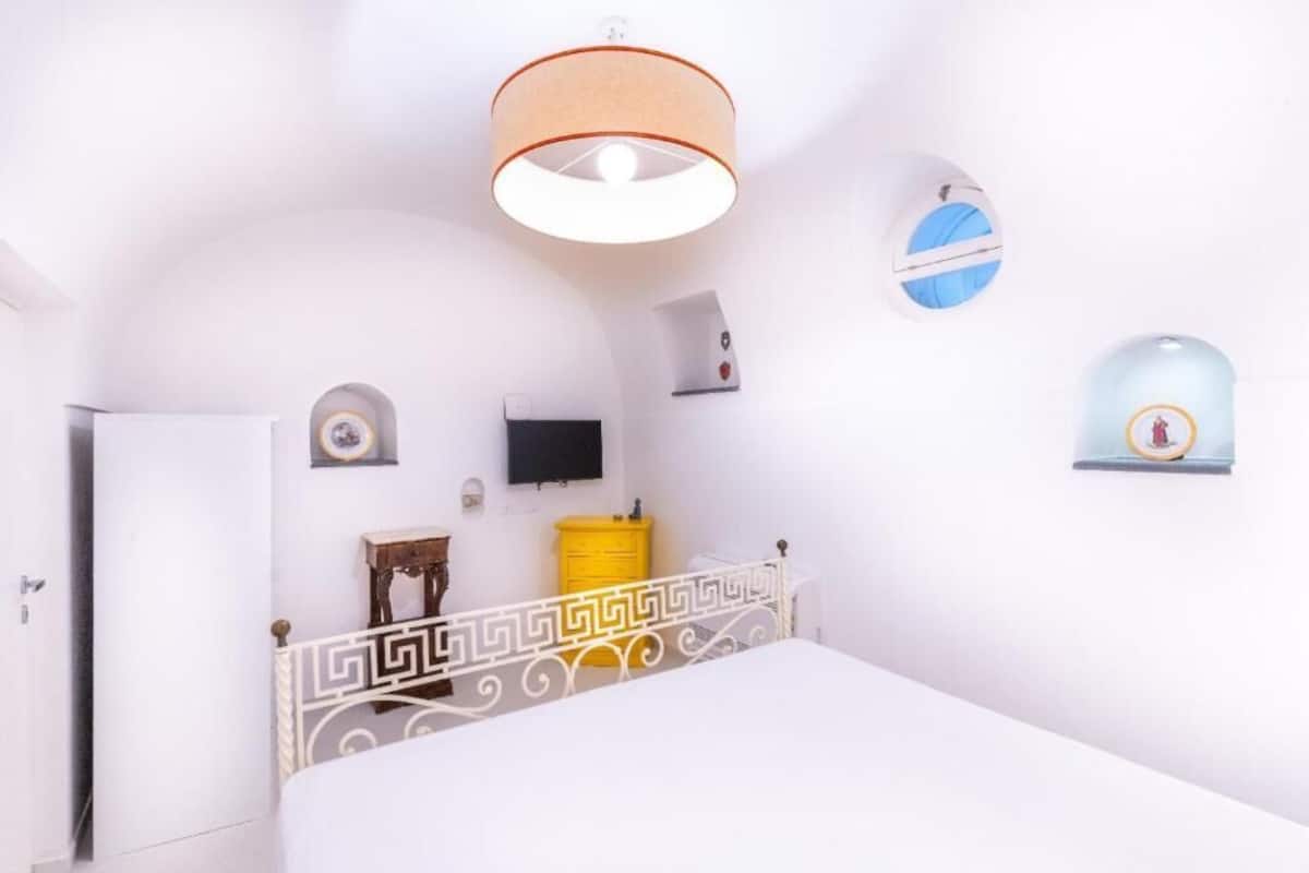 A white room with a bed, a television, a yellow trash bin, and a door.