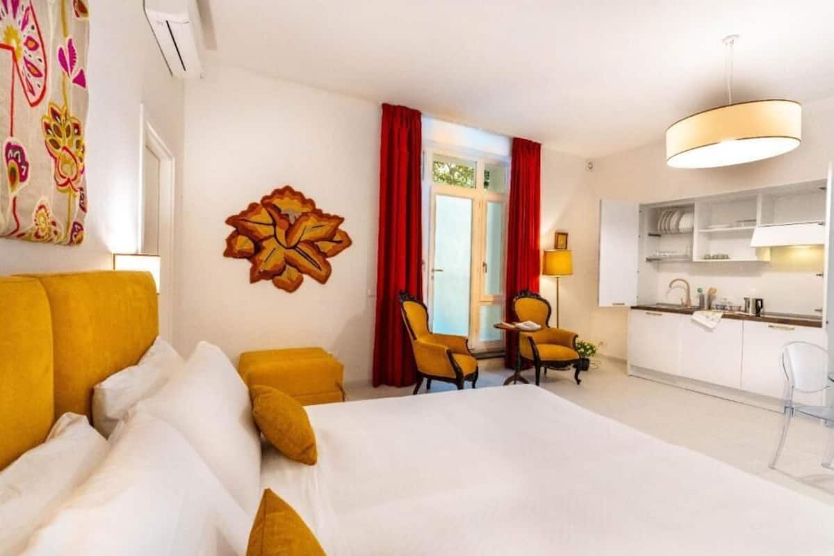 A hotel room with a bed, a desk, two chairs, a kitchen area, and a large window with red curtains.