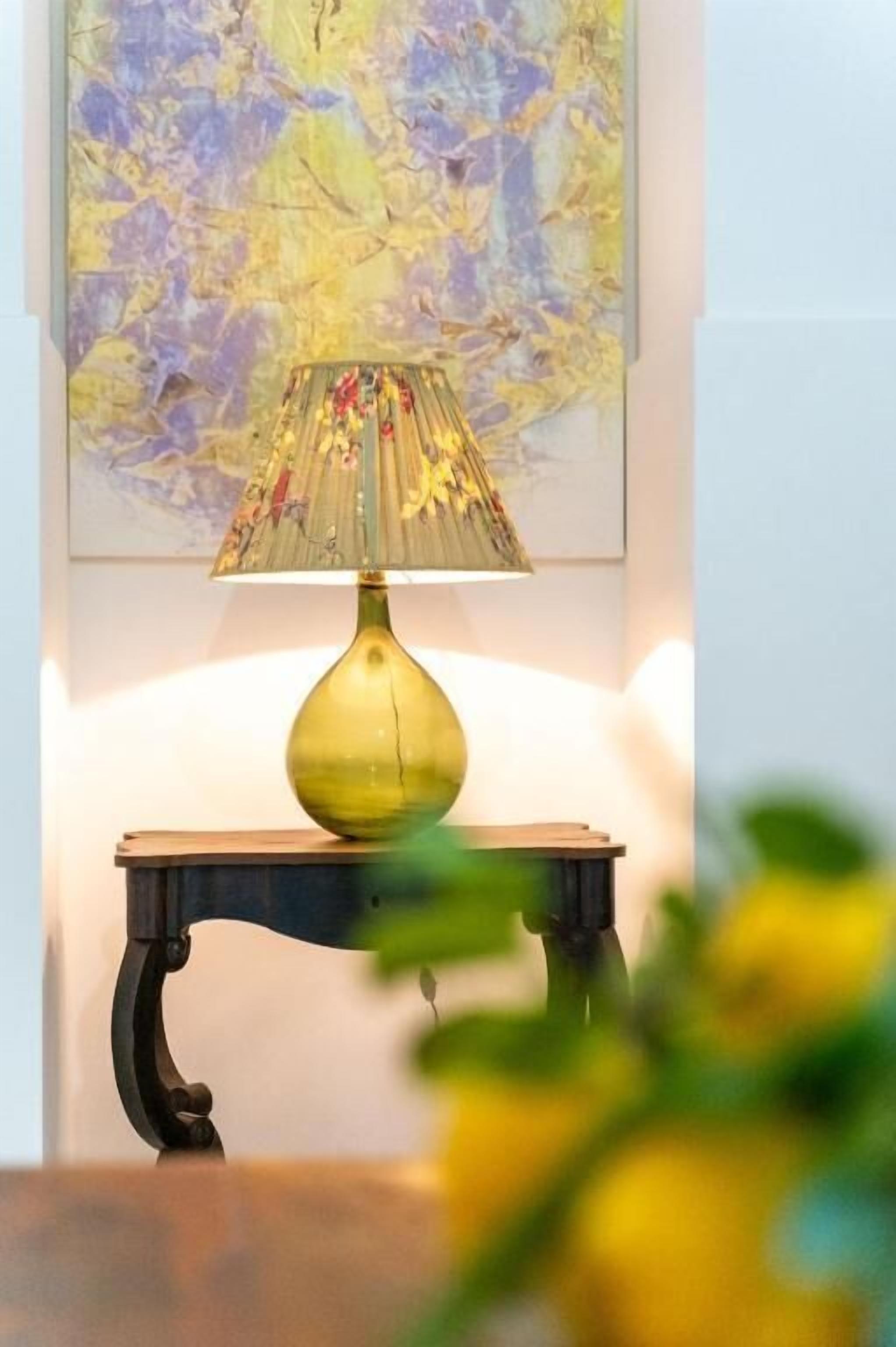 Una mesa con una lámpara, un cuadro en la pared y una planta desenfocada al frente.
