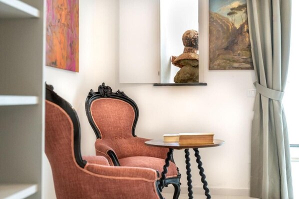 A room with a red upholstered armchair, a small round table with a book, and artwork on the walls. - Dimora Filangieri (Massa Lubrense)