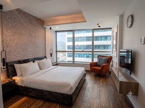Ein modernes Hotelzimmer mit einem großen Bett, einer Sitzecke, einem Fernseher und Blick auf Gebäude durch große Fenster.