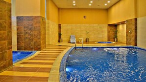 Uma grande piscina interior com chão e paredes revestidas de azulejos, uma pequena área de estar com uma toalha, e um relógio na parede.