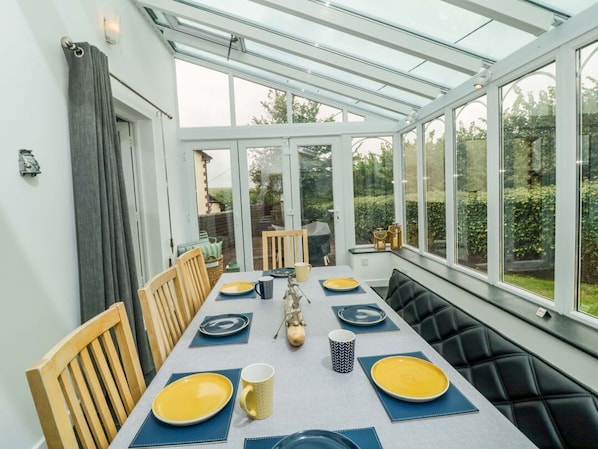 Un espace repas aménagé dans une véranda avec un toit en verre, des chaises en bois et une table dressée pour quatre personnes.