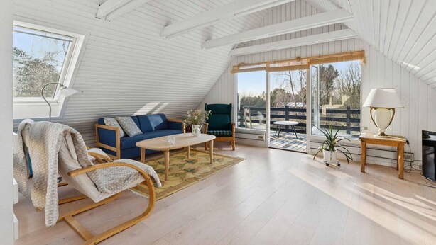 A spacious attic room with a high ceiling, a blue sofa, a wooden rocking chair, a small table, and a large window offering a view of the outdoors. - 4 Star Holiday Home in Humble (Sydlangeland)