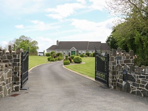 A gated driveway leading to a house with a stone wall on one side and a well-maintained lawn with trimmed bushes.