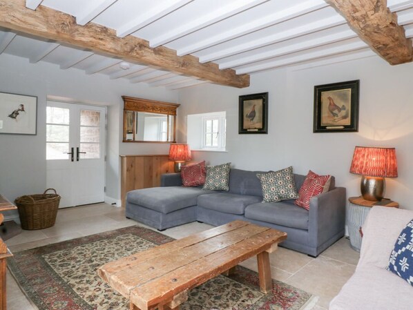 Ein Wohnzimmer mit einer Eckcouch, einem Holztisch, einem gemusterten Teppich und einer Decke mit sichtbaren Holzbalken.
