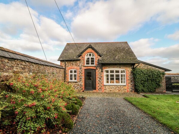 A stone house with a gravel driveway, a black door, and a small garden with red flowers.