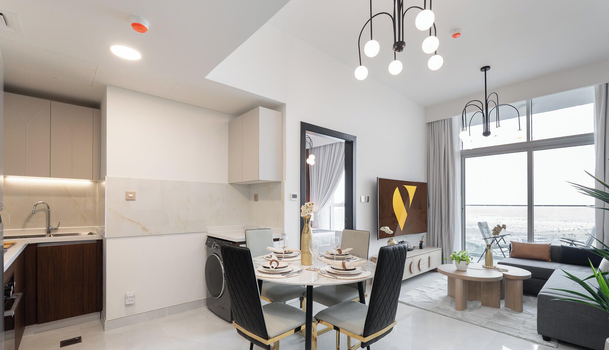 A modern kitchen with a built-in washing machine, a dining area with a round table and chairs, and a large window offering a view.