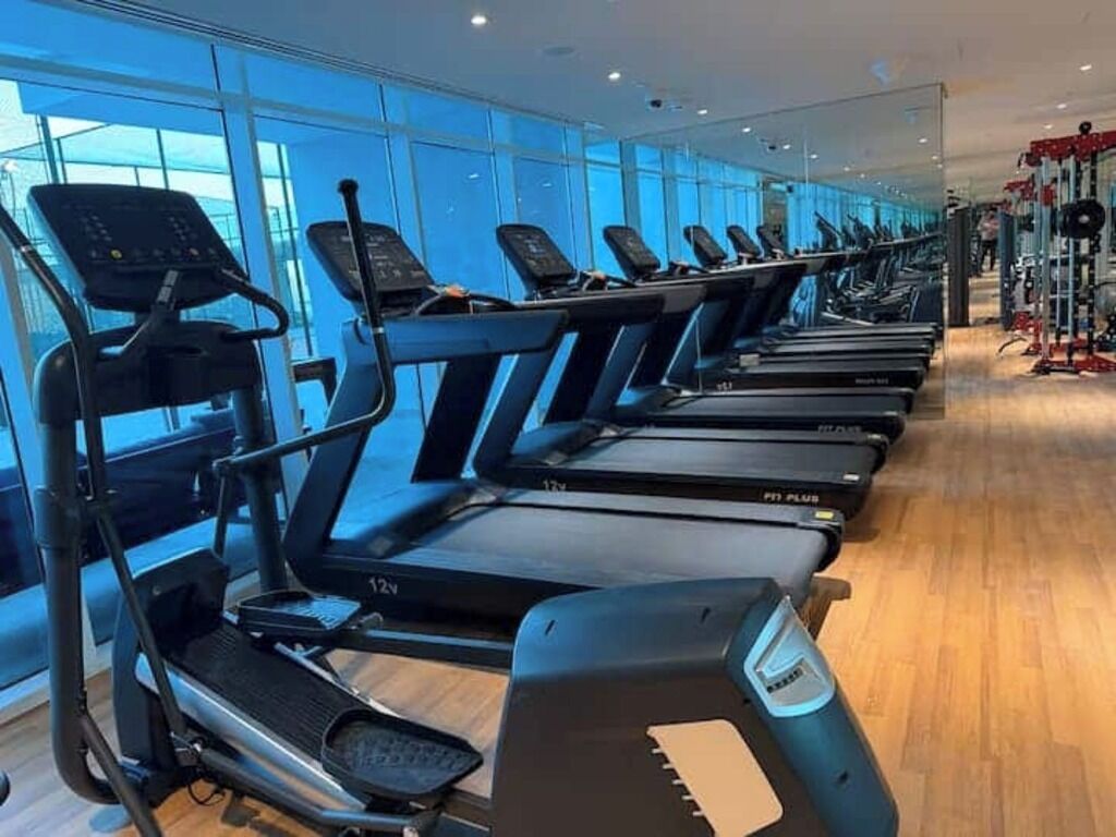 Une rangée de tapis de course dans une salle de sport dotée de grandes fenêtres et d’un plancher en bois.