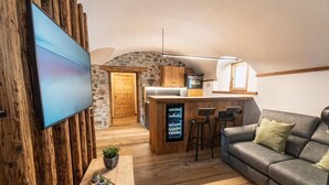 A cozy living area with a TV, a sofa, a coffee table with plants, a kitchen with a stone wall, and a bar area. - Giuly's Home Luxury Bormio – Alpine Comfort and Wellness (Bormio)