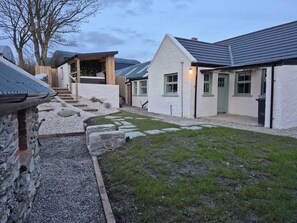 A stone building with a gravel pathway, a wooden structure, and a white house with green windows. - Annie’s Farm Cottage Retreat (Londonderry)