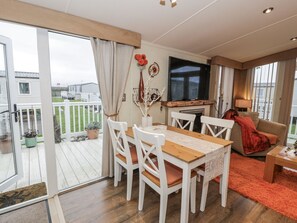 A spacious living area with a wooden dining table, white chairs, a flat-screen TV, and a cozy seating area with a red blanket. - Beachside Retreat in Towyn (Abergele)