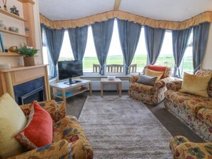 A living room with a fireplace, a television, a sofa, and a window with curtains. - Cae Carreg, Cemaes Bay (Cemaes Bay)