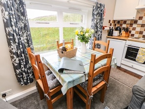 A dining area with a table set for four, wooden chairs, a vase with flowers, and a kitchen in the background. - Hobson's Cottage - Pet-Friendly Retreat (Richmond)