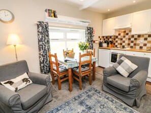 A cozy living room with a dining table, two grey sofas, a blue rug, and a kitchen area with white cabinets and a tiled backsplash. - Hobson's Cottage - Pet-Friendly Retreat (Richmond)