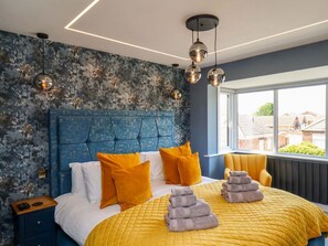 Ein Schlafzimmer mit blauem Kopfteil, einer gelben Bettdecke und Blick durch ein großes Fenster nach draußen.