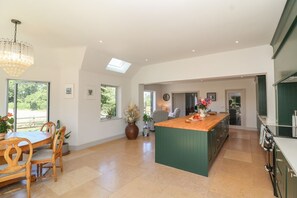 A spacious kitchen with a large island, a dining area with a round table, and a view of greenery outside. - Leafield Cottage, Brockworth (Gloucester)