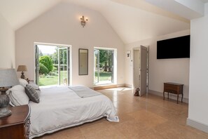 A bedroom with a large bed, a TV mounted on the wall, and a sliding glass door leading to a garden. - Leafield Cottage, Brockworth (Gloucester)