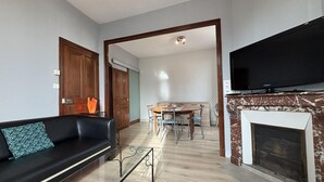 Ein Wohnzimmer mit schwarzem Sofa, einem Esstisch mit Stühlen, einem Kamin und einem darüber montierten Fernseher.