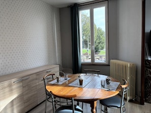 A room with a wooden dining table, metal chairs, a bench, a radiator, and a window with a view of greenery.