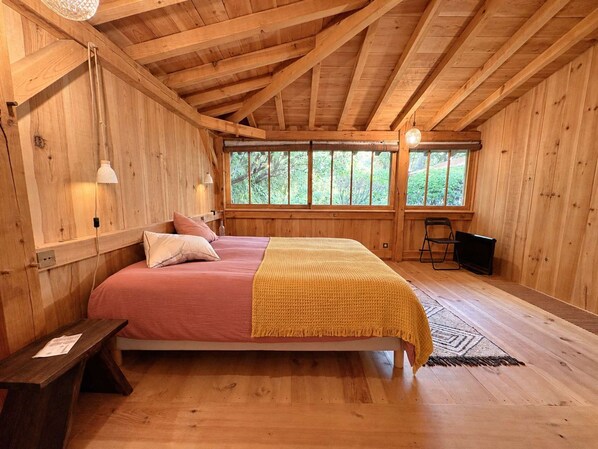 Un dormitori de cabana de fusta amb un llit, taulells laterals, una finestra i una taula petita.