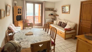 Ein gemütliches Zimmer mit Esstisch, Holzstühlen, einer Couch, einem Fernseher und einem Balkon mit Aussicht.