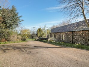 Ein Feldweg führt zu einem Gebäude aus Stein mit geneigtem Dach, umgeben von Bäumen und Vegetation.