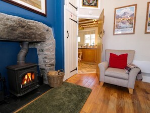 A cozy room with a stone fireplace, a grey sofa with a red pillow, and a kitchen visible through an open door. - 1 Bryn Ysgol, Betws-Y-Coed (Betws-y-Coed)