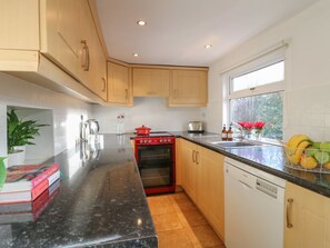 A kitchen with wooden cabinets, a red stove, a dishwasher, and a window with a view of trees. - 2 Gladstone Avenue, Melton Mowbray (Melton Mowbray)