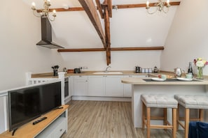 A modern kitchen with a TV, wooden beams, a central island, and bar stools. - The Old Dairy, Nettleton (Chippenham)