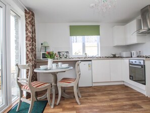 A kitchen with white cabinets, a dining table with chairs, a window with a green valance, and a built-in oven. - Sunset Cottage, Tintagel (Tintagel)