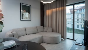 Una sala de estar moderna con un sofá modular, una mesa de comedor con platos y un vaso, un cuadro en la pared y vistas de un edificio a través de un ventanal.
