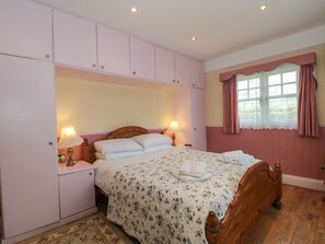 A bedroom with a wooden bed, a nightstand with a lamp, a wardrobe, and a window with curtains. - Rose Cottage, Swanage, Dorset. (Swanage)
