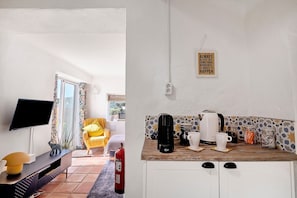 Una habitación con televisión, sofá, sillón y una zona de cocina con cafetera, tazas y frascos.