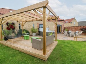 A wooden pergola with seating, a stone building, and a grassy yard.