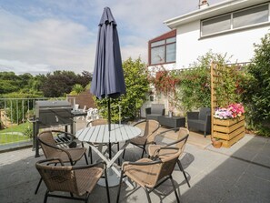 An outdoor patio with a round table, four wicker chairs, a grill, and a wooden planter with flowers.