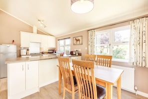 Une cuisine avec des meubles blancs, une table à manger entourée de chaises en bois, un luminaire suspendu et une fenêtre avec des rideaux.