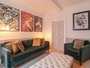 A living room with a dark green sofa, a matching armchair, a tufted ottoman, and three framed botanical prints on the wall.