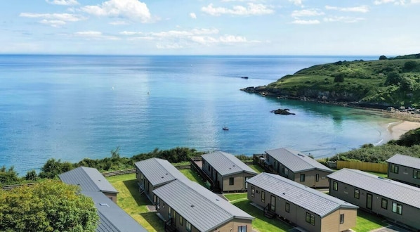 Udsigt over kysten med en række moderne, etplans hytter med flade tage, placeret på en græsmark med fri udsigt til havet og et bakket landskab i baggrunden.